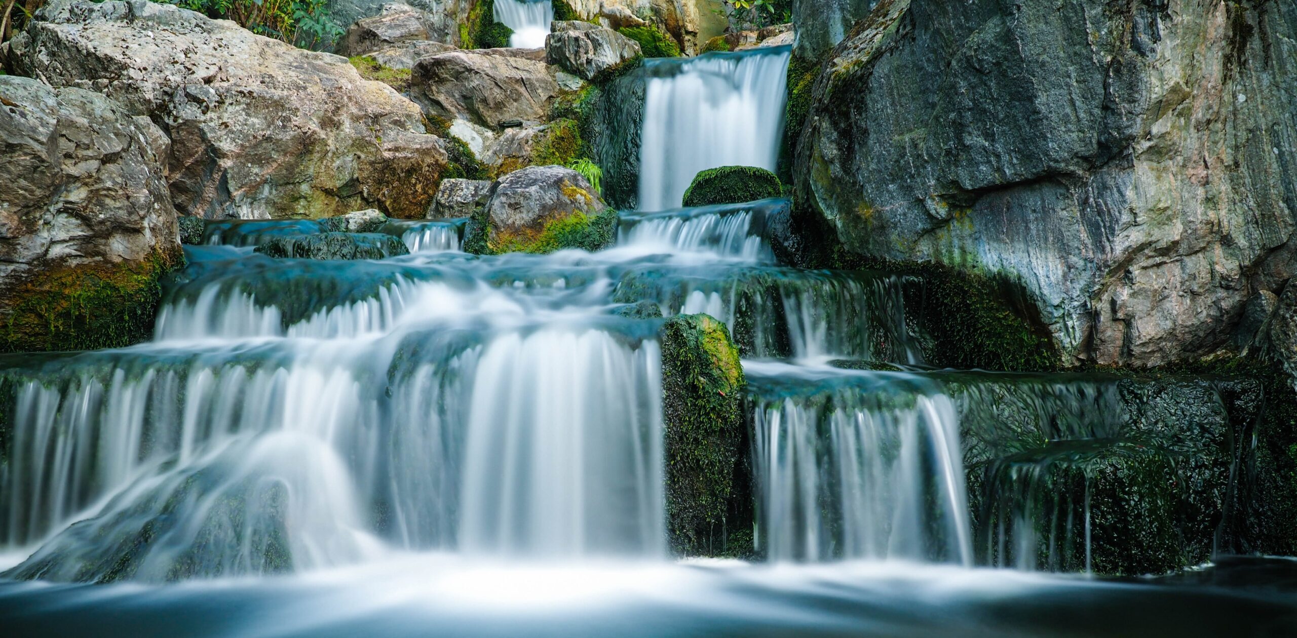 Slow-shutter image of a multi-tiered waterfall, with extensive moss growth around the lower tiers and grayish-brown rocks catching direct sunlight near the top.