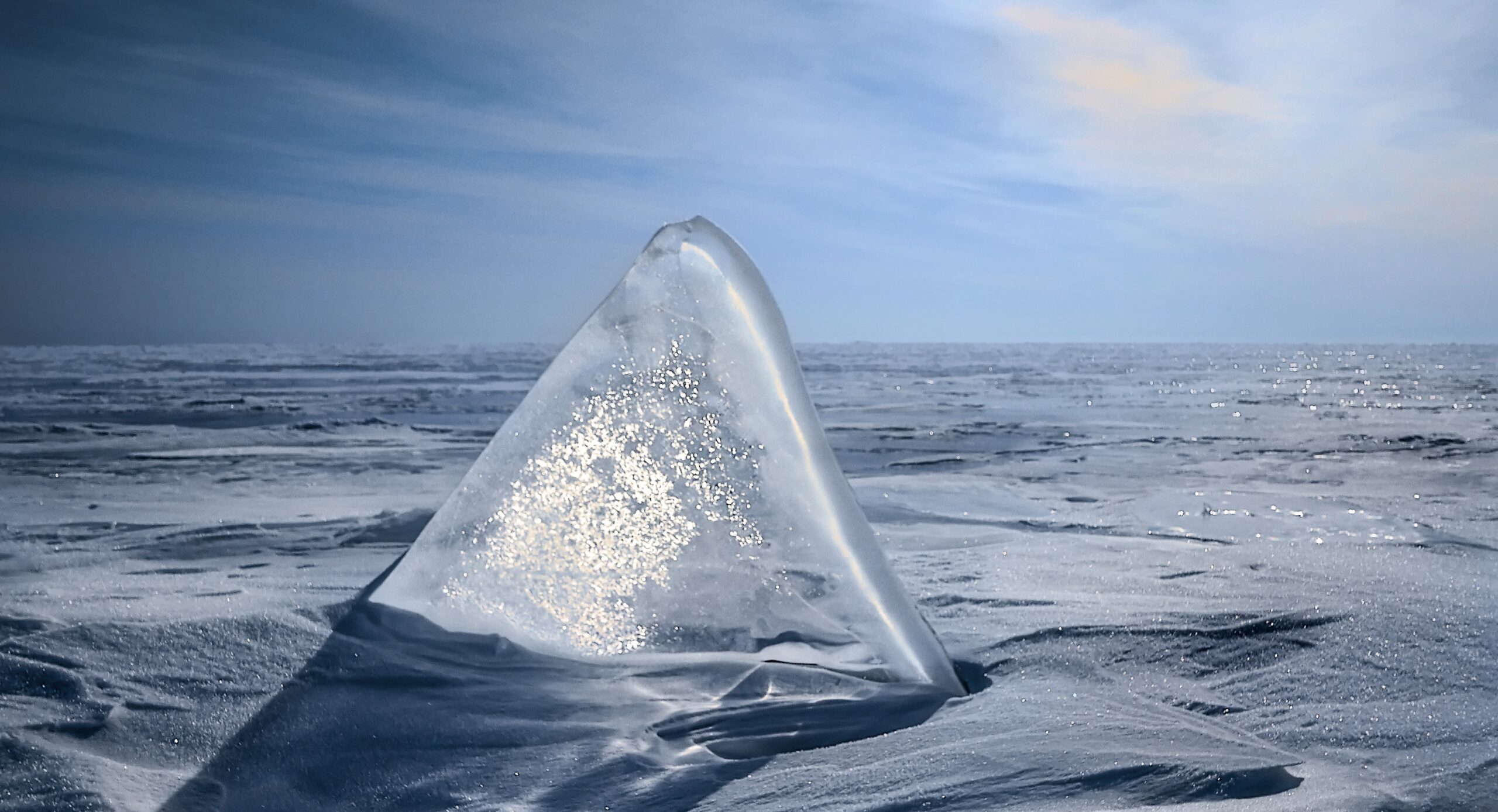 Ice in the shape of a shark fin emerges from a snow field, sunlight filtering through wisps of clouds in the background. Is ice a sort of historical water calendar?