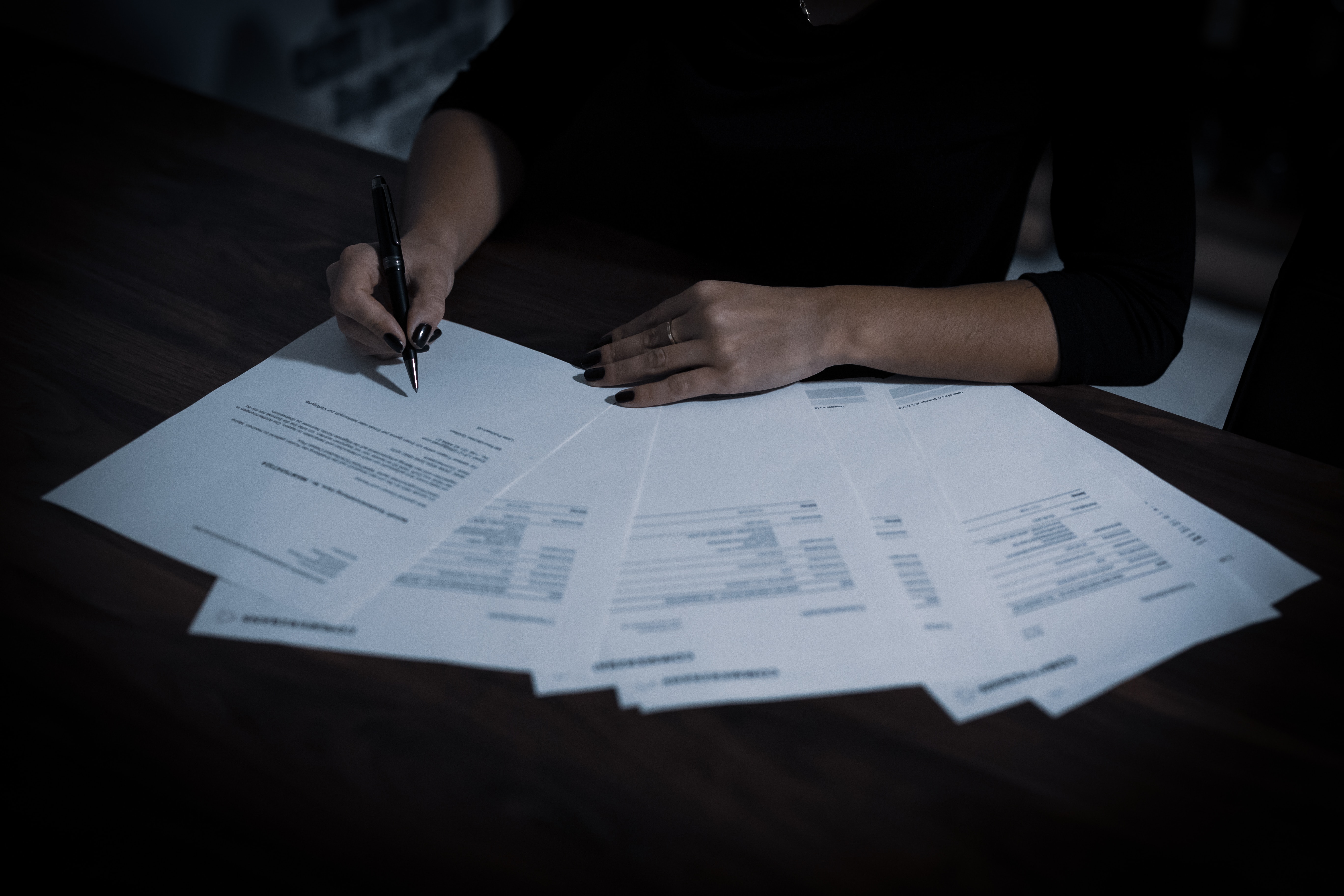 A white woman sits at a table with several papers spread out in front of her. She holds a pen and appears to be signing one of the papers.