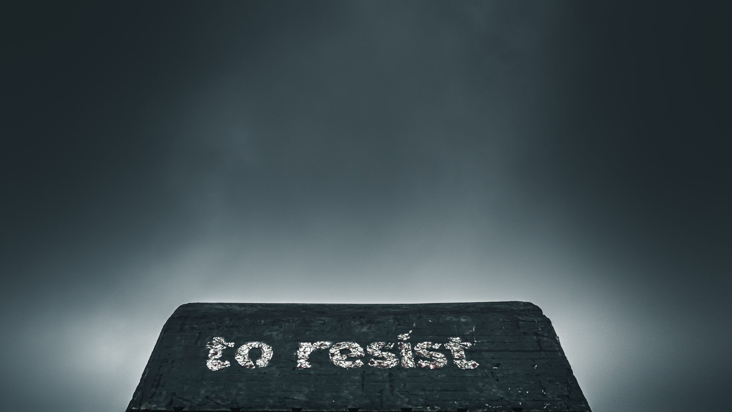 Against a gray sky, a darker gray brick wall rises with the words "to resist" painted on in white block letters.